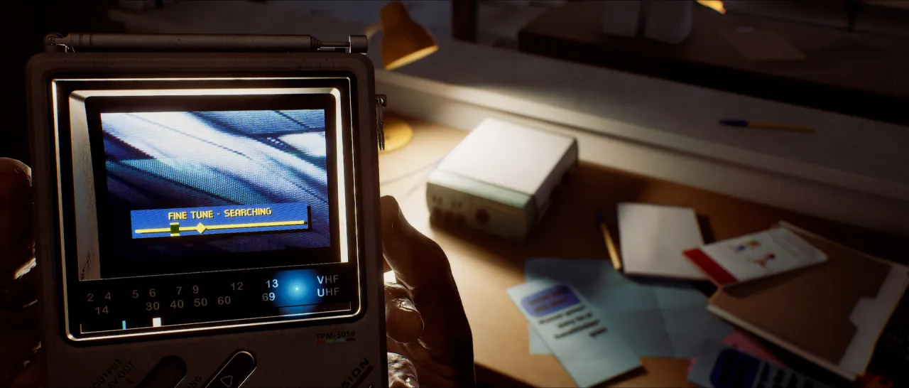First-person view of a handheld television with a fine-tune bar reading searching, above a desk of papers.